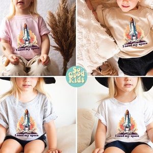 K&ouml;nnte beinhalten: Vier T-Shirts f&uuml;r Kinder mit einem Space-Shuttle-Motiv und dem Text "Ich brauche meinen Platz". Die Shirts sind in verschiedenen Farben: rosa, beige, grau und wei&szlig;.