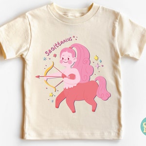 May include: A cream coloured t-shirt with a pink Sagittarius zodiac sign illustration. The illustration shows a pink centaur with a bow and arrow. The text "Sagittarius" is written in pink above the illustration.