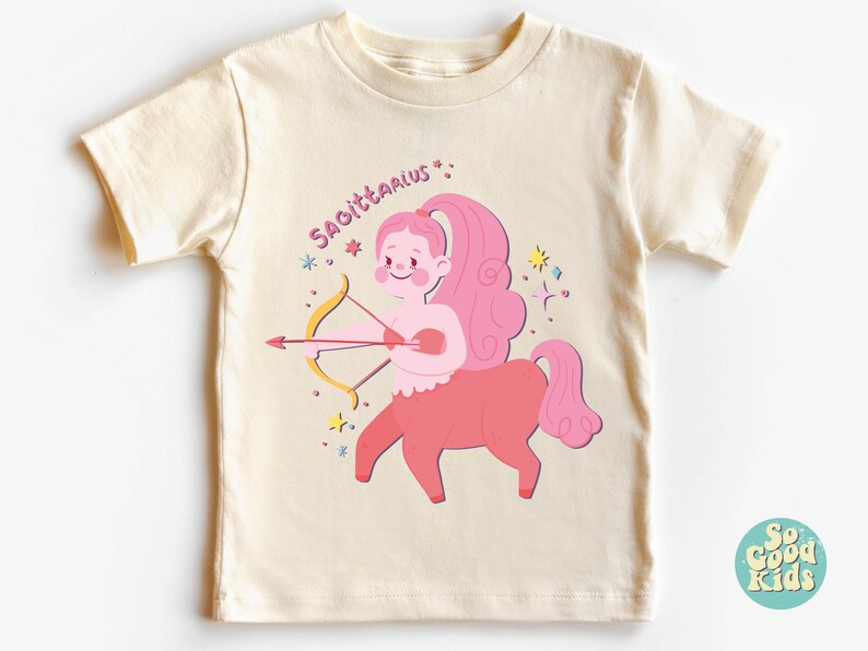 May include: A cream coloured t-shirt with a pink Sagittarius zodiac sign illustration. The illustration shows a pink centaur with a bow and arrow. The text "Sagittarius" is written in pink above the illustration.