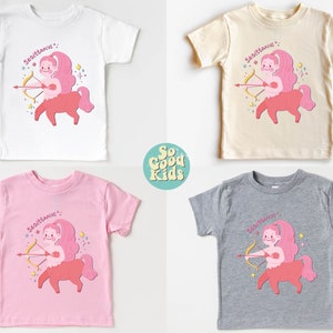 May include: Four toddler-sized t-shirts featuring a pink and red illustration of a Sagittarius zodiac sign with the text "Sagittarius" above the illustration. The shirts are in white, beige, pink, and grey.