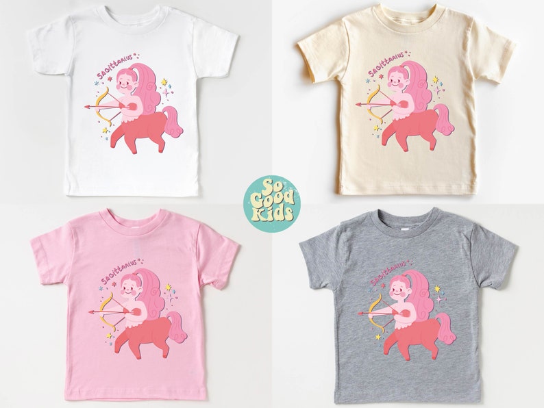 May include: Four toddler-sized t-shirts featuring a pink and red illustration of a Sagittarius zodiac sign with the text "Sagittarius" above the illustration. The shirts are in white, beige, pink, and grey.