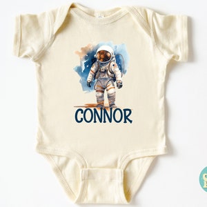 May include: A cream-colored baby onesie with a watercolor illustration of an astronaut on the moon. The onesie has the name "CONNOR" printed in blue below the astronaut.