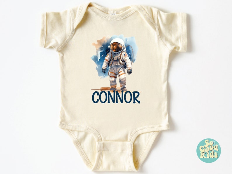 May include: A cream-colored baby onesie with a watercolor illustration of an astronaut on the moon. The onesie has the name "CONNOR" printed in blue below the astronaut.