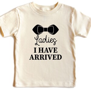 May include: A cream colored t-shirt with a black bow tie graphic and the text "Ladies I Have Arrived".