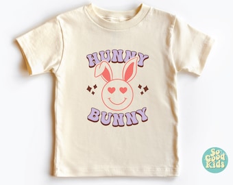 Peut inclure: Un t-shirt à manches courtes de couleur crème avec un lapin rose aux yeux en forme de cœur et le texte "Hunny Bunny" en lettres violettes et brunes.