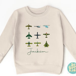 May include: A cream-colored sweatshirt featuring various airplane illustrations in green, blue, and brown. The name "Jackson" is written in a cursive font below the aircraft. The sweatshirt has a crew neck and long sleeves.