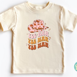 K&ouml;nnte beinhalten: Ein cremefarbenes T-Shirt mit einem rosa und braunen Cowboyhut mit Kuhmuster und dem dreimal wiederholten Text "Yee Haw" in Orange, Gelb und Braun.