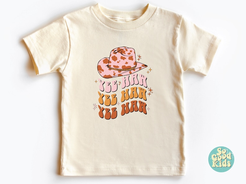 K&ouml;nnte beinhalten: Ein cremefarbenes T-Shirt mit einem rosa und braunen Cowboyhut mit Kuhmuster und dem dreimal wiederholten Text "Yee Haw" in Orange, Gelb und Braun.