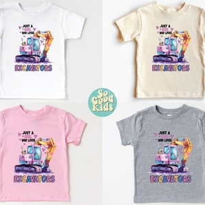 May include: Four children's t-shirts in white, beige, pink, and gray, each featuring a colorful excavator graphic. The text reads "Just a girl who loves excavators". The shirts are from the "So Good Kids" collection.