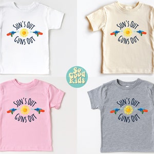 May include: Four children's t-shirts in white, beige, pink, and gray, each featuring the text "SUN'S OUT GUNS OUT" with a sun and water guns graphic. The shirts are short-sleeved.