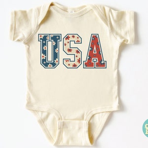 K&ouml;nnte beinhalten: Ein cremefarbener Baby-Strampler mit kurzen &Auml;rmeln zeigt eine Grafik im Vintage-Stil des Wortes "USA" in Blau und Rot mit einem floralen Muster.