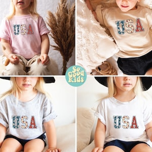 K&ouml;nnte beinhalten: Vier Kinder tragen T-Shirts in verschiedenen Farben mit dem Wort "USA" darauf gedruckt im Vintage-Stil mit roten, wei&szlig;en und blauen Blumenmustern. Die Shirts sind in Pink, Beige, Grau und Wei&szlig; erh&auml;ltlich.