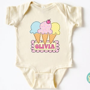 May include: A cream-colored baby onesie with a graphic of three ice cream cones in blue, pink, and yellow with a cherry on top. The ice cream cones are arranged in a triangle shape and are sitting on top of a pink and white striped banner with the name "Olivia" written in pink letters.