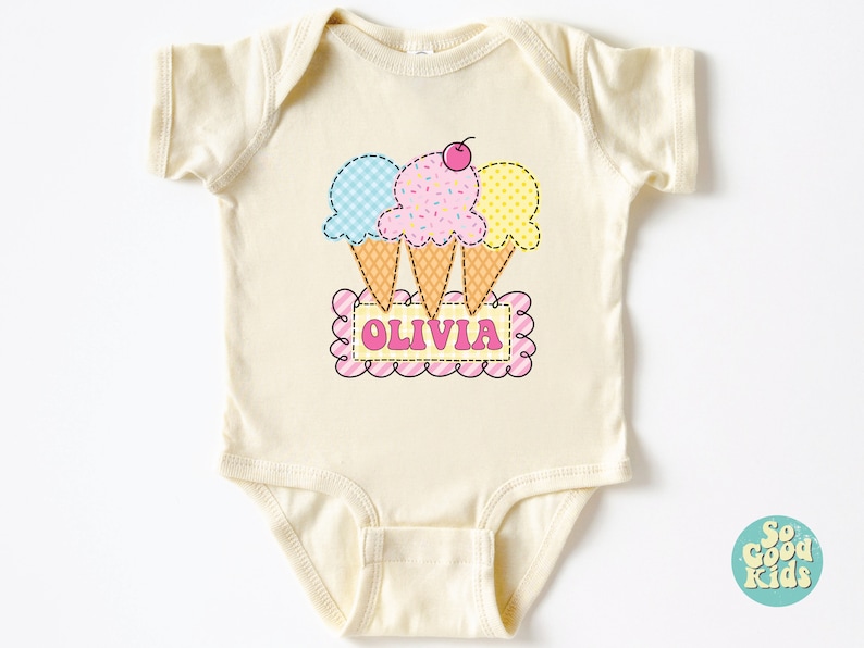 May include: A cream-colored baby onesie with a graphic of three ice cream cones in blue, pink, and yellow with a cherry on top. The ice cream cones are arranged in a triangle shape and are sitting on top of a pink and white striped banner with the name "Olivia" written in pink letters.
