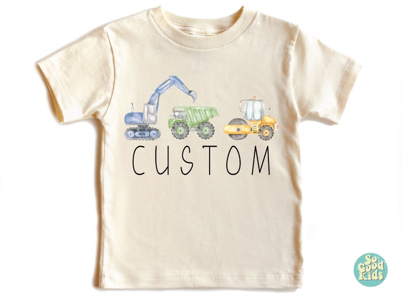May include: A light beige t-shirt with a blue, green, and yellow watercolor illustration of construction vehicles. The text "CUSTOM" is printed in black below the vehicles.