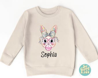 May include: A beige sweatshirt with a colorful bunny wearing pink heart-shaped glasses and blowing a bubble. The bunny has a pink bow on its head. The text "Sophia" is printed below the bunny.