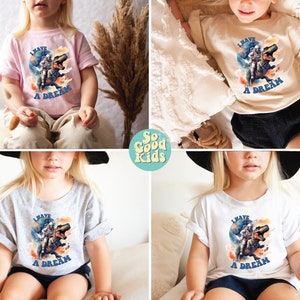 Pode incluir: Quatro crian&ccedil;as est&atilde;o vestindo camisetas de cores diferentes com uma estampa gr&aacute;fica de um astronauta montando um dinossauro. O texto nas camisetas diz "Eu tenho um sonho".