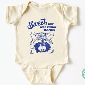 Op de afbeelding: Een cr&egrave;mekleurige baby romper met een blauwe wasbeer afbeelding en de tekst "Sweet, but will throw hands".