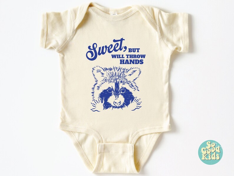 Op de afbeelding: Een cr&egrave;mekleurige baby romper met een blauwe wasbeer afbeelding en de tekst "Sweet, but will throw hands".
