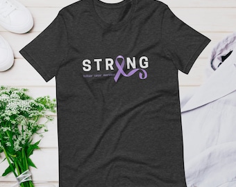 Puede incluir: Una camiseta gris oscuro con la palabra "STRONG" en letras blancas y una cinta morada. El texto "testicular cancer awareness" está impreso debajo de la cinta.