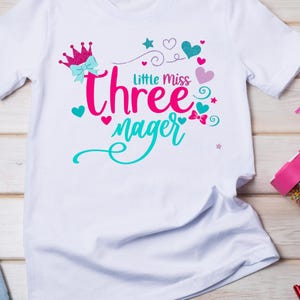 May include: White t-shirt with a pink crown, a pink bow, and the text "Little Miss three" in pink and "nager" in turquoise.