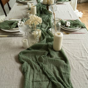 Puede incluir: Una mesa decorada con un camino de mesa y servilletas verde salvia. La mesa está adornada con platos blancos, cristalería transparente y velas blancas. Flores secas y bolas de algodón añaden un toque natural a la elegante decoración.
