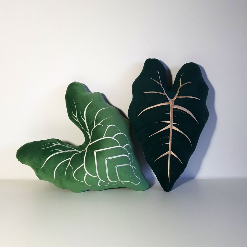 May include: Two green velvet throw pillows shaped like leaves. One pillow has a white outline of a leaf, the other has a gold outline.