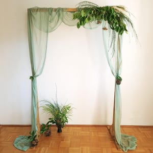 May include: A wooden archway decorated with green foliage and draped with light green sheer fabric. The archway is standing on a wood floor.