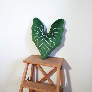 May include: A green velvet pillow shaped like a heart with white veins. The pillow is sitting on a wooden step stool.
