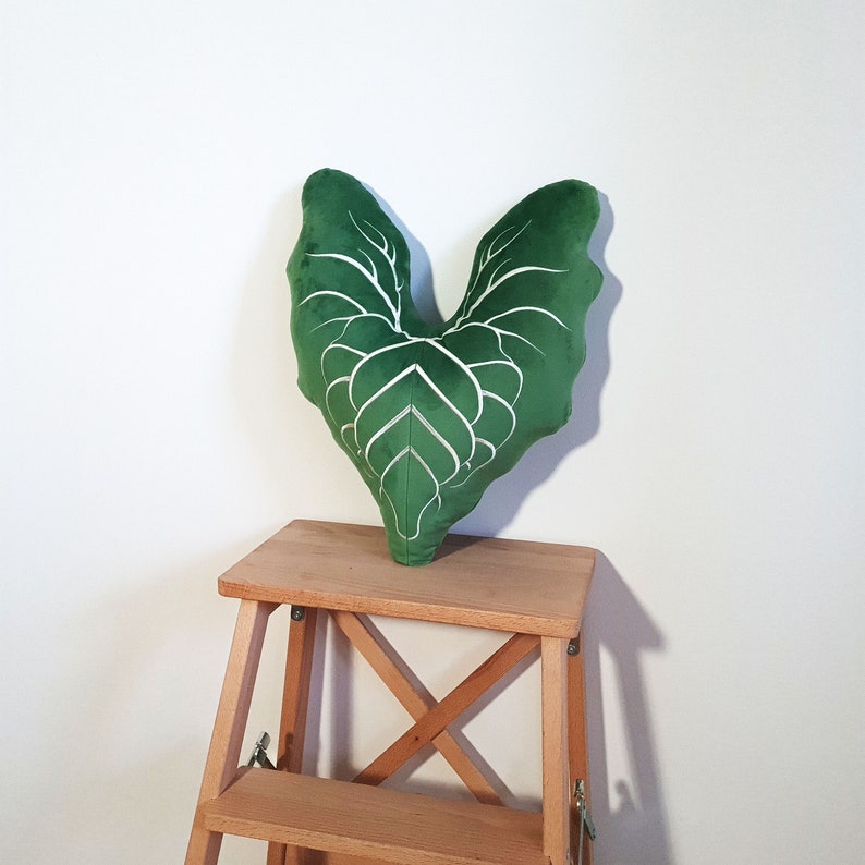May include: A green velvet pillow shaped like a leaf with white veins. The pillow is sitting on a wooden step stool.