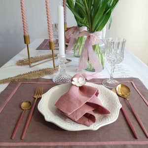 May include: A blush pink linen placemat with a white, embossed, round dinner plate, a pink linen napkin with a white flower napkin ring, gold flatware, and a gold spoon. The place setting is on a table with a white tablecloth and a bouquet of green tulips in a glass vase.