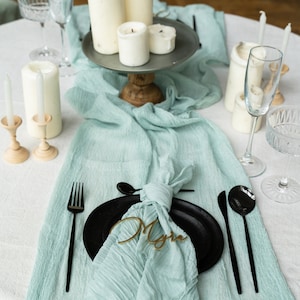 May include: A light blue linen napkin tied around a black plate with a gold name card that says "Myra". The plate is set on a white tablecloth with a light blue runner. There are white candles and candle holders on the table.