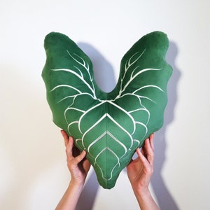 May include: A green velvet pillow shaped like a leaf with white embroidered veins. The pillow is held by two hands.