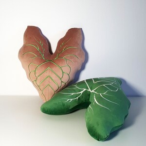 May include: Two decorative pillows shaped like leaves. The larger pillow is a heart shape with a brown velvet fabric and green embroidery. The smaller pillow is a green leaf shape with white embroidery.
