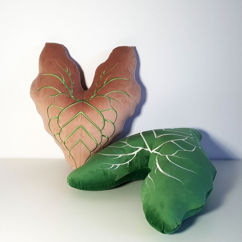May include: Two decorative pillows shaped like leaves. The larger pillow is a heart shape with a brown velvet fabric and green embroidery. The smaller pillow is a green leaf shape with white embroidery.