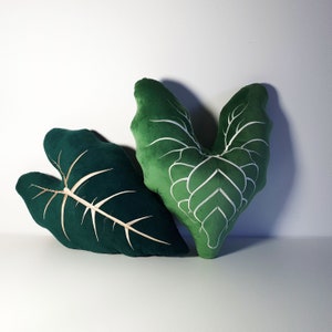 May include: Two green velvet throw pillows shaped like leaves. One pillow has a white embroidered design, while the other is solid green.