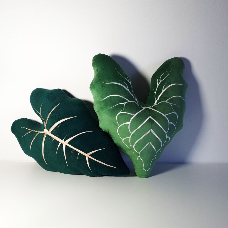 May include: Two green velvet throw pillows shaped like leaves. One pillow has a white embroidered design, while the other is solid green.
