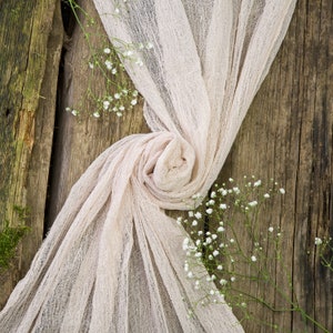 Puede incluir: Una delicada tela de color rosa pálido drapeada sobre una superficie de madera rústica. La tela se reúne en el centro, creando una forma suave y fluida. Pequeñas flores blancas se esparcen alrededor de la tela, añadiendo un toque de elegancia.