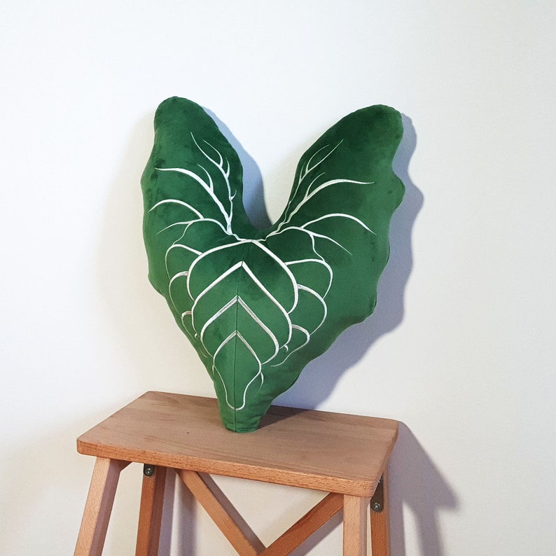 May include: A green heart-shaped pillow with white leaf veins embroidered on the front. The pillow is sitting on a wooden stool.
