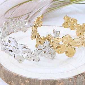 Könnte beinhalten: Zwei Armbänder mit Blumendesign, eines silberfarben und eines goldfarben. Jedes Armband hat mehrere blütenförmige Ornamente. Die Armbänder werden auf einer hellen Holzoberfläche präsentiert.