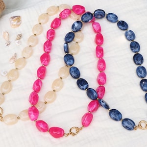 Puede incluir: Tres collares con cuentas ovaladas en color crema, rosa brillante y azul jaspeado. Cada collar está formado por cuentas de resina pulida separadas por pequeños espaciadores metálicos dorados y un cierre de palanca de tono dorado.