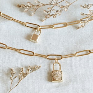 Peut inclure: Deux colliers en chaîne dorée avec des breloques en forme de cadenas. Le collier du haut a un breloque de cadenas simple, tandis que le collier du bas a un breloque de cadenas avec un design en forme de cœur recouvert de petits strass.