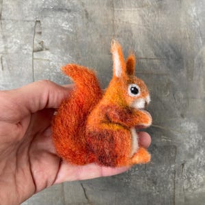 Könnte beinhalten: Eine handgefertigte Eichhörnchenfigur aus Filz in Orange- und Brauntönen. Das Eichhörnchen hat einen buschigen Schwanz, kleine Ohren und schwarze Augen. Die Figur wird in einer Hand gehalten, um ihre Größe und Details zu zeigen. Ein bezauberndes Dekorationsobjekt.