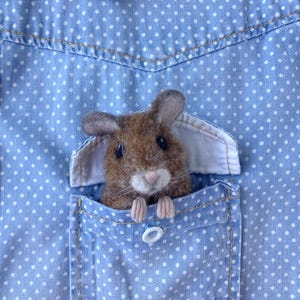 Może przedstawiać: Brązowa filcowa mysz wygląda z kieszeni niebieskiej koszuli w kropki. Mysz ma małe czarne oczy, różowy nos i maleńkie łapki. Kieszeń koszuli jest szczegółowo wykończona białymi szwami i białym guzikiem.