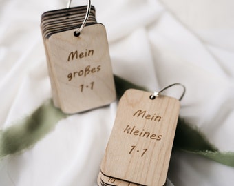 Puede incluir: Dos juegos de tarjetas de memoria de madera con anillos plateados. El juego superior dice "Mein großes 1-1" y el juego inferior dice "Mein kleines 7-7".