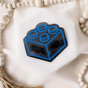 Könnte beinhalten: Ein blauer und schwarzer Aufkleber mit einem klassischen Bausteinspielzeug. Der Aufkleber befindet sich auf einer weißen Oberfläche.