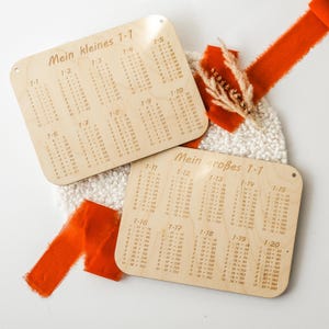 Könnte beinhalten: Zwei Holztafeln mit darauf gedruckten Multiplikationstabellen. Die kleinere Tafel trägt den Titel "Mein kleines 1-7" und zeigt Multiplikationstabellen von 1-10. Die größere Tafel trägt den Titel "Mein großes 1-7" und zeigt Multiplikationstabellen von 1-20.