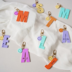 Könnte beinhalten: Bunte Schlüsselanhänger aus Acryl in verschiedenen Farben, jeweils in Form eines Buchstabens des Alphabets. Jeder Schlüsselanhänger hat einen goldfarbenen Verschluss und einen Smiley-Anhänger. Namen sind auf die Buchstaben gedruckt. Die Buchstaben sind orange, gelb, blau, lila und türkis.