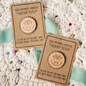 Könnte beinhalten: Zwei braune Karten mit deutschem Text und einer Holzmonster-Münze. Die Karten liegen auf einer weißen, gewebten Oberfläche mit einem hellgrünen Band. Kleine Smiley-Anstecknadeln sind verstreut.