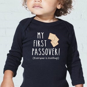 May include: A dark blue baby bodysuit with the text "MY FIRST PASSOVER!" in white. The design includes an illustration of matzah and the phrase "(Everyone is kvelling!)". The bodysuit is designed for a baby or toddler.